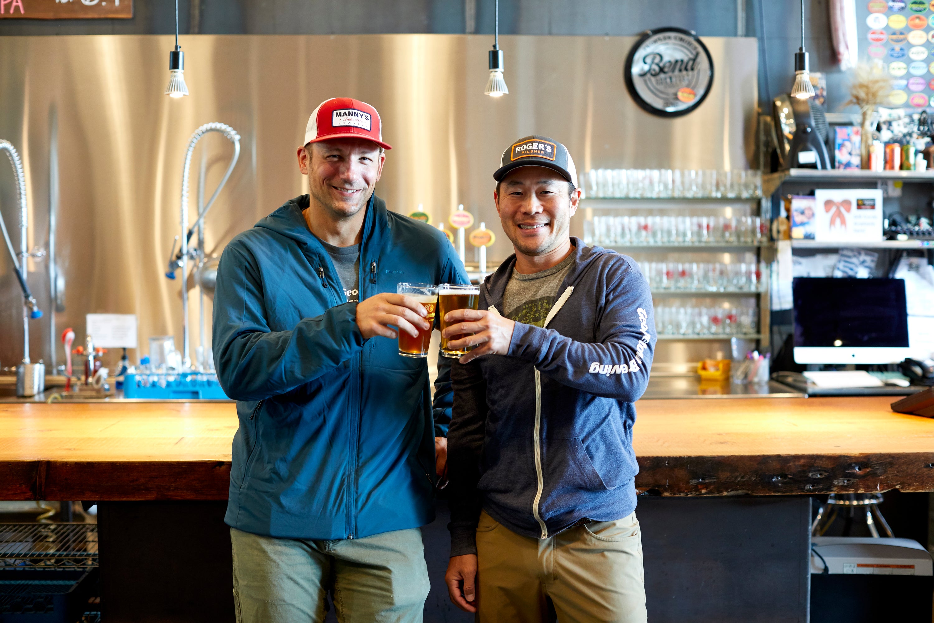 Two men standing in a bar or brewery, holding drinks and smiling.
